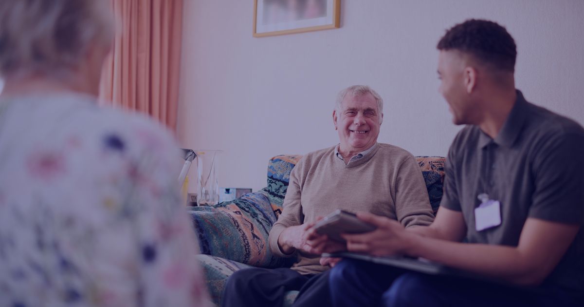 An older man and a younger man sit on a sofa, smiling and talking, with a woman in the foreground.