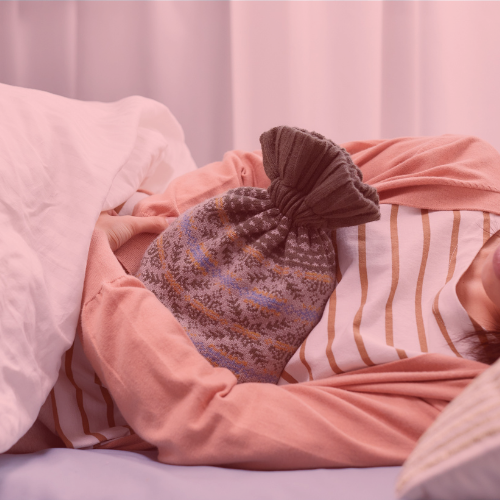 Person lying in bed, partially covered by a blanket, holding a patterned hot water bottle against their stomach