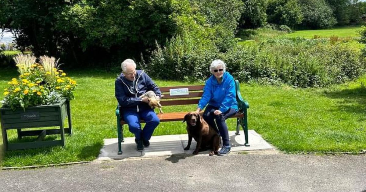 a man and his wife sat happily on one of Bury's Age Friendly benches in the park with their two dogs