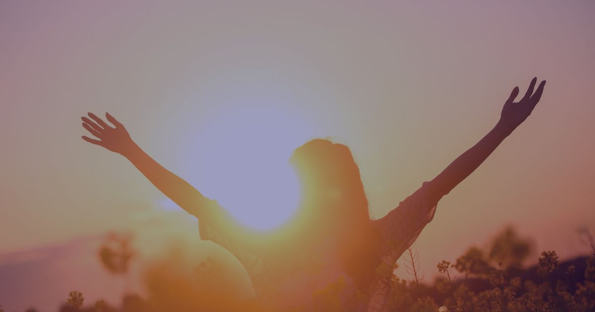 a woman looking out in to the distance, with her arms outstretched and the sun setting in the distance