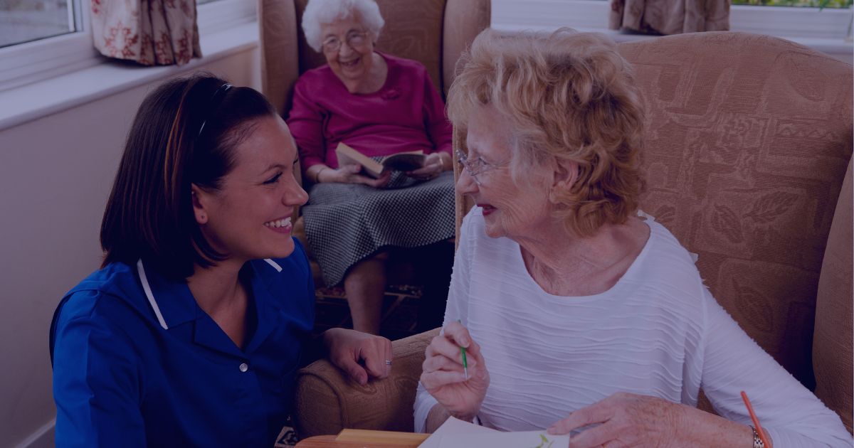 A young female carer in a blue uniform smiles at an elderly woman who is painting; another elderly woman reads a book in the background.