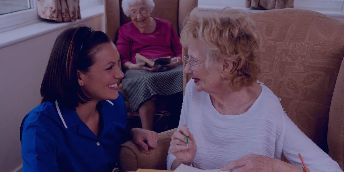 A young female carer in a blue uniform smiles at an elderly woman who is painting; another elderly woman reads a book in the background.