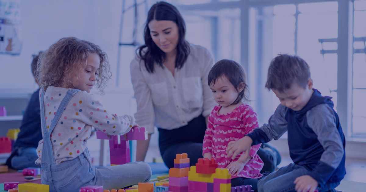 A group of young children and an adult playing together with large colorful building blocks in a bright, modern classroom.