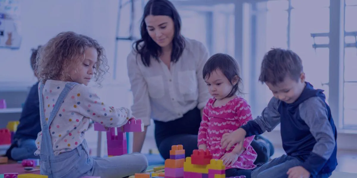 A group of young children and an adult playing together with large colorful building blocks in a bright, modern classroom.