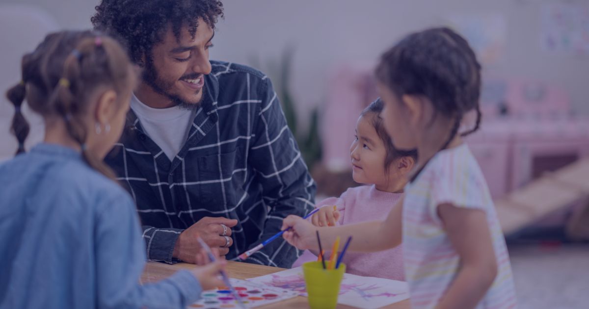 a man sat at a table with 3 young children, smiling and helping them to paint a picture