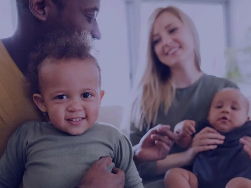 A smiling family sitting together indoors: a man holding a toddler and a woman holding a baby.