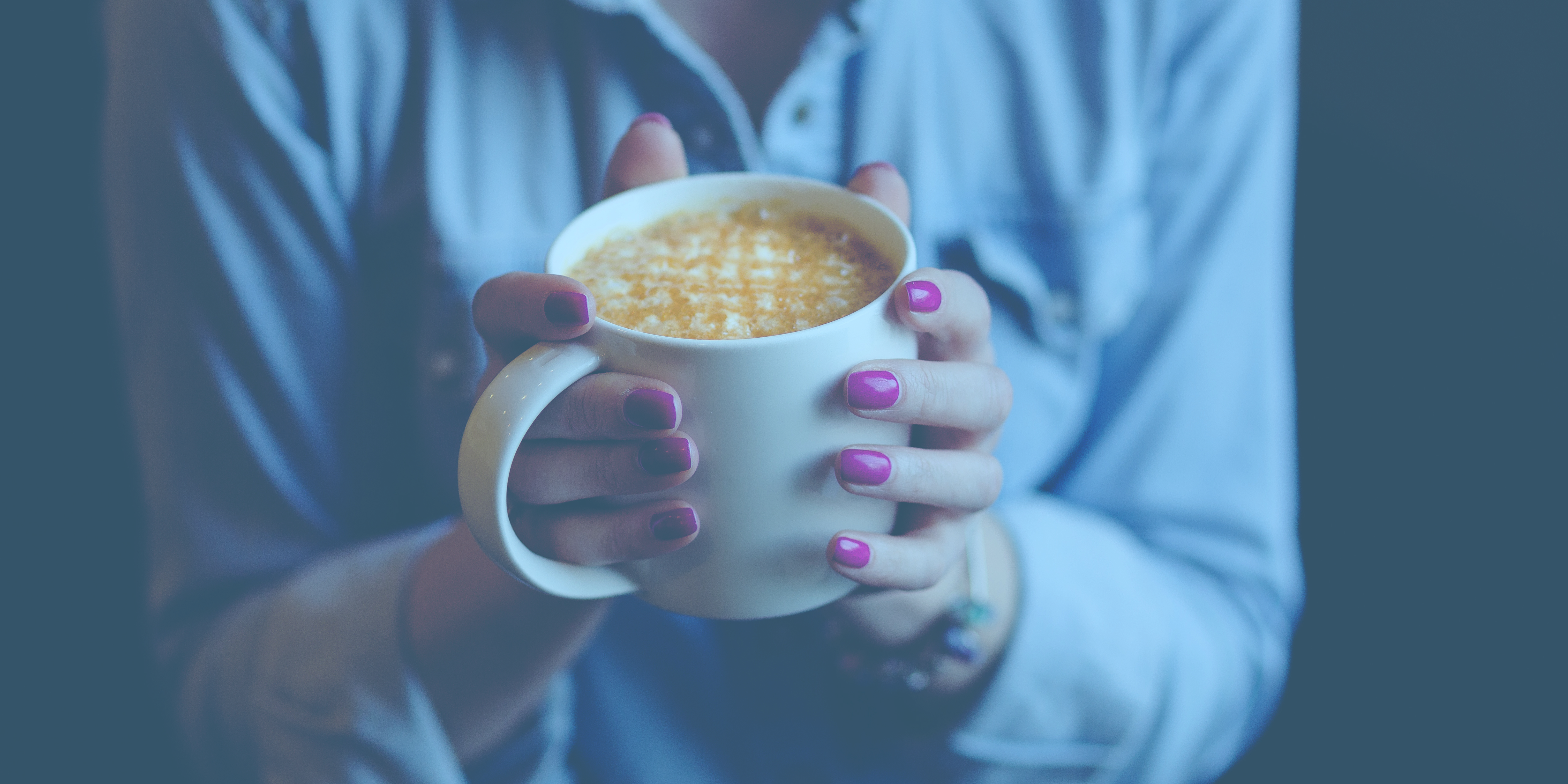 Person holding a white mug filled with a frothy hot drink, wearing a light blue shirt and pink nail polish