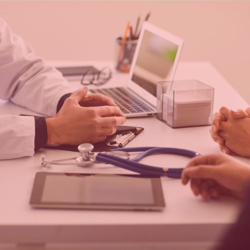 A doctor's hands gesturing during a consultation at a desk with a laptop, tablet, stethoscope, and a patient's hands resting.