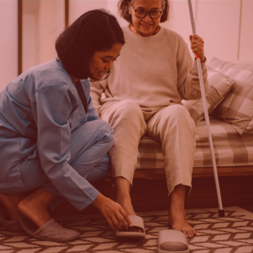 caregiver in blue scrubs helps an older woman sitting on bed put on her slippers; the woman holds a white cane