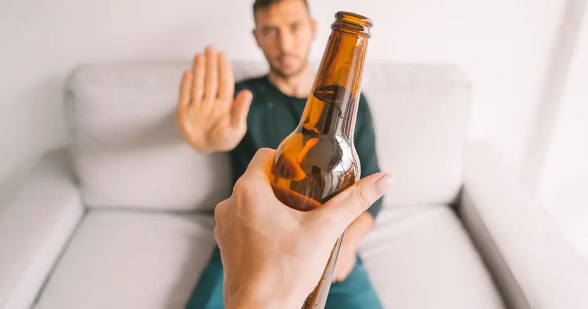 a hand holding out a bottle of beer offering it to a man sat on a sofa. The man appears to decline by holding his hand out to say stop
