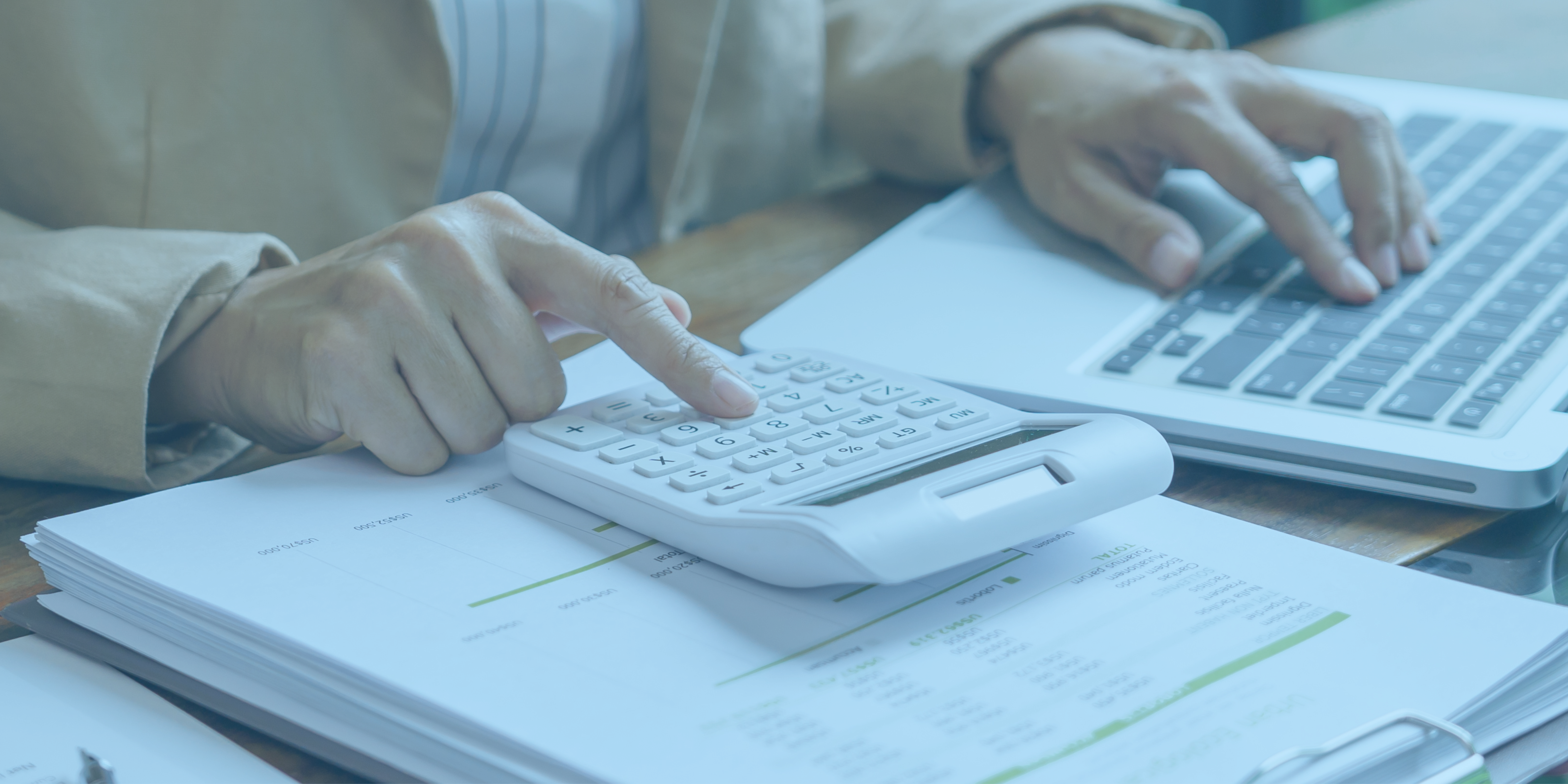 Close-up of hands using a calculator on top of financial documents, with a laptop keyboard in the background