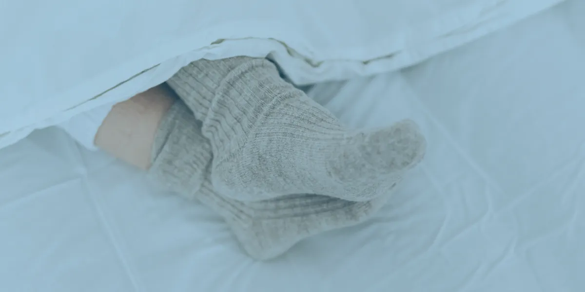 Close-up of feet wearing thick grey socks under a white blanket on a bed