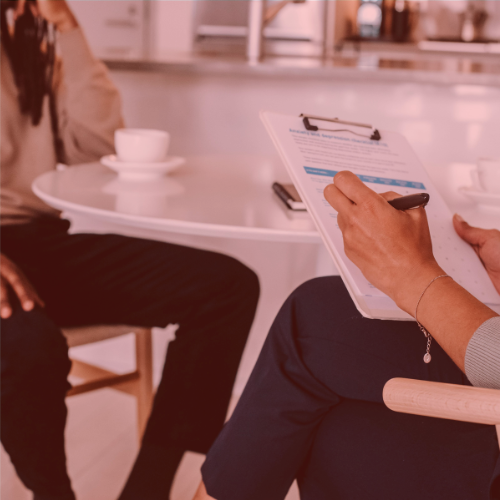 Two individuals sit at a round table in a kitchen, one holding a clipboard and pen, and the other with a coffee cup