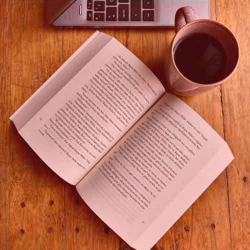 Open book on wooden table beside a laptop and a brown mug of black coffee.