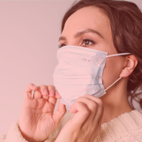 Woman adjusting a blue surgical mask over her nose and mouth, wearing a cream sweater