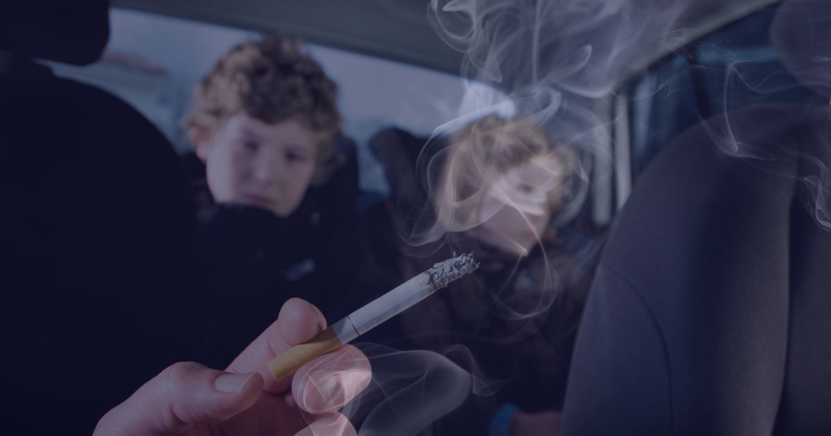 Two young children sat in the rear of a vehicle, whilst a hand holds a lit, cigarette with smoke circulating around the car