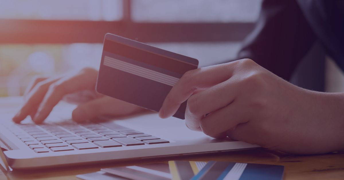 A person holds a credit card while typing on a laptop, with several other cards on the table.