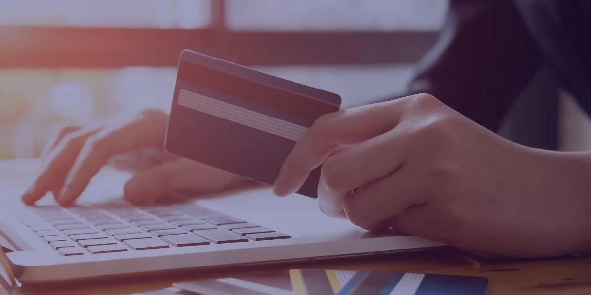 A person holds a credit card while typing on a laptop, with several other cards on the table.