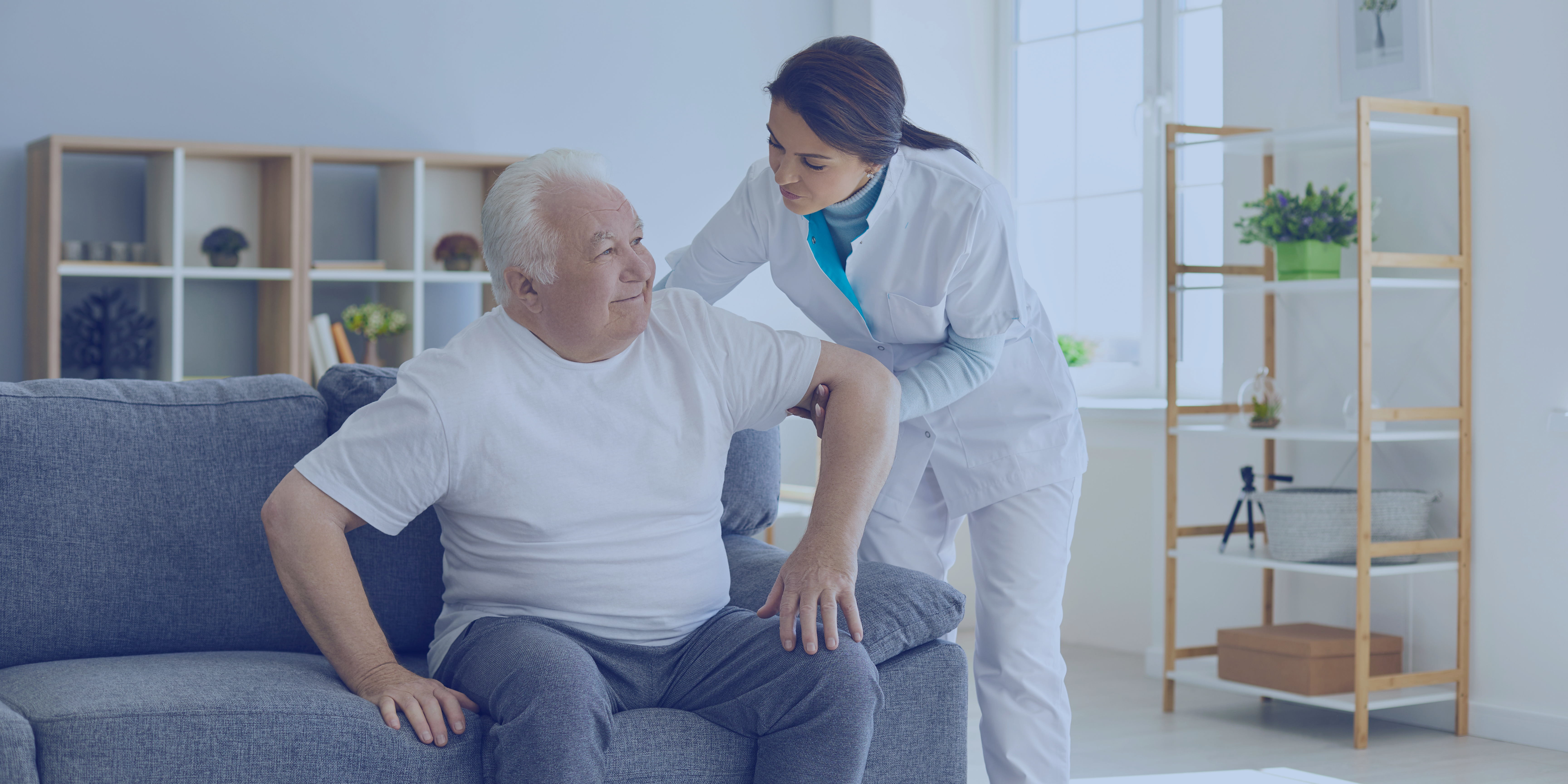 A healthcare professional assists an elderly man sitting on a grey couch in a well-lit room.