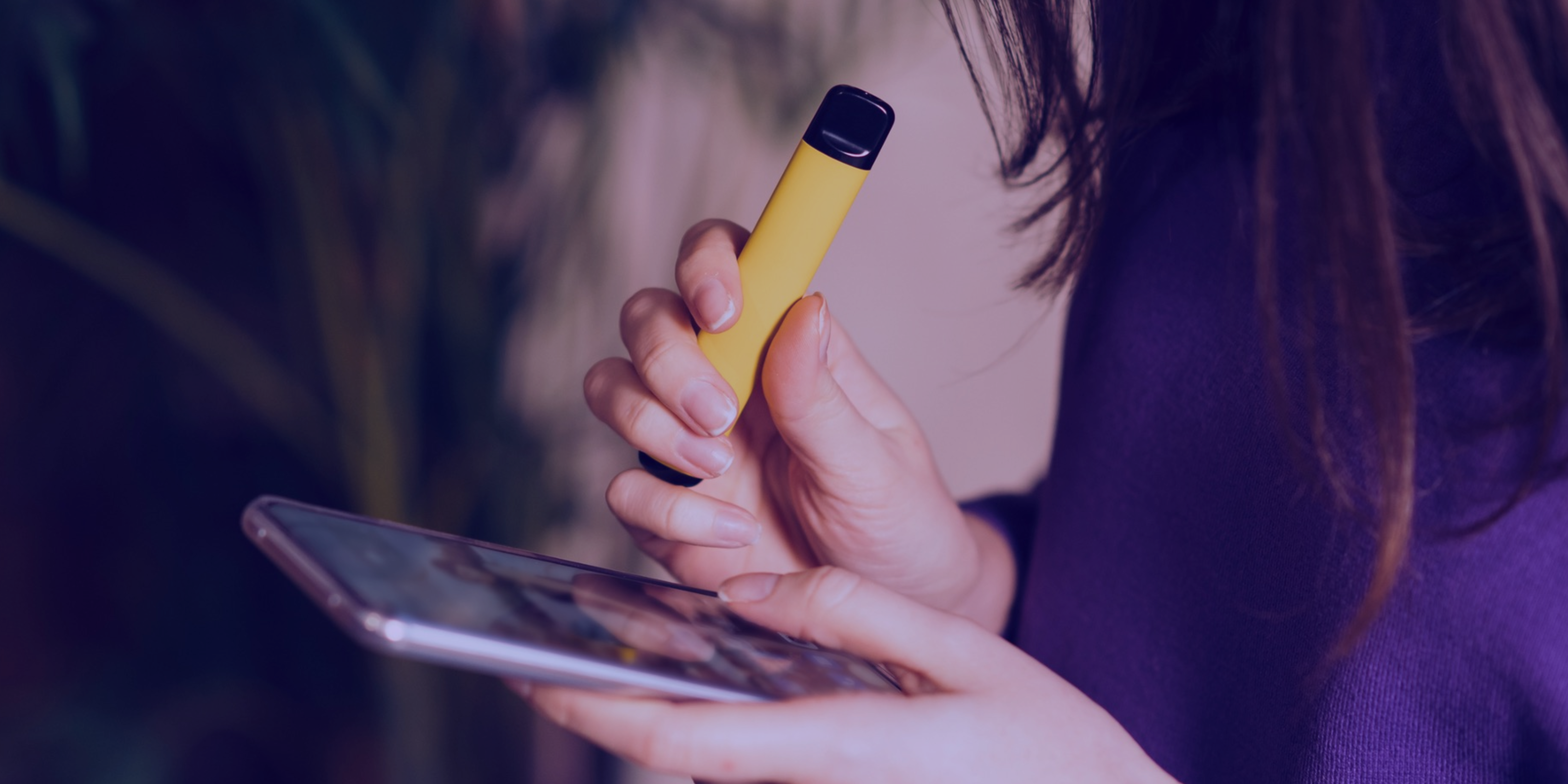 Close-up of person holding yellow disposable vape in one hand and smartphone in the other.