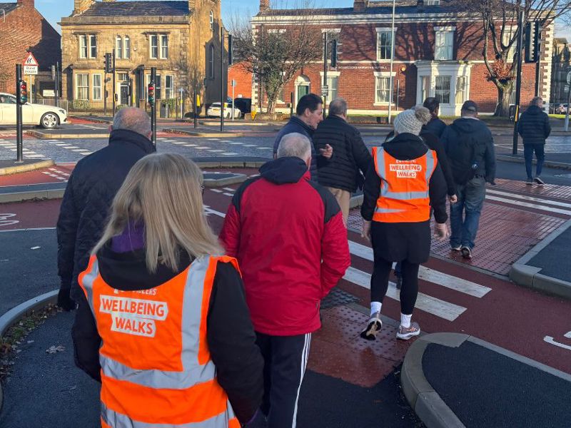 a group of people walking in Bury town centre, alongside volunteers wearing orange vests printed with the words 'wellbeing walks'