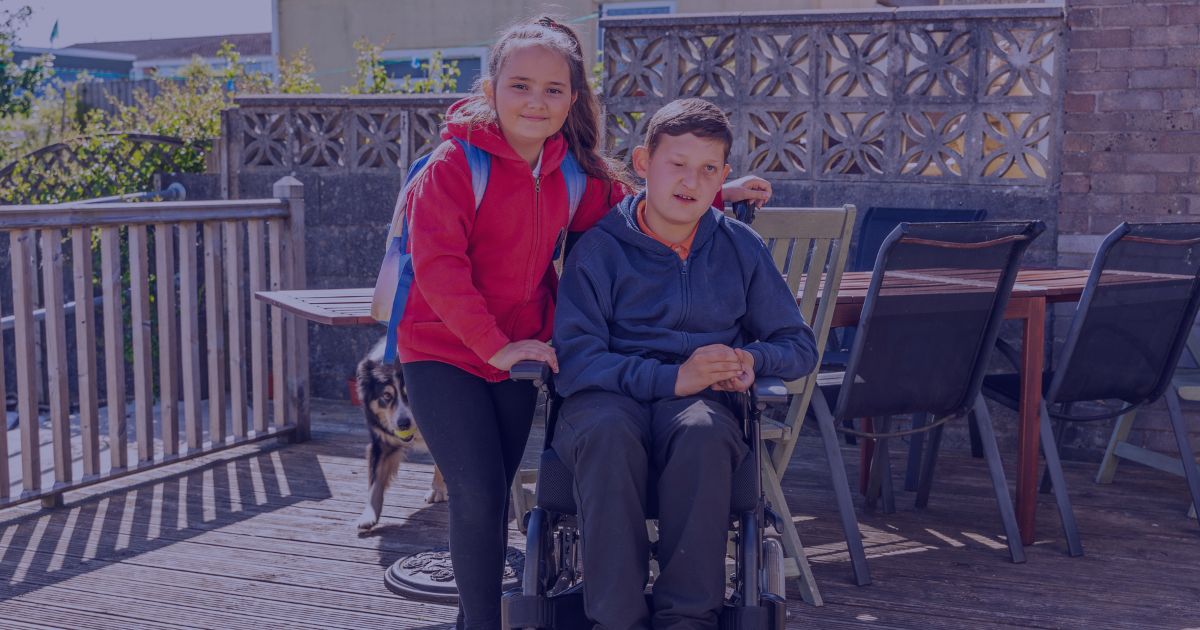 A young girl in a red hoodie standing next to a boy in a wheelchair on a wooden deck, with a dog walking behind them and outdoor furniture in the background.