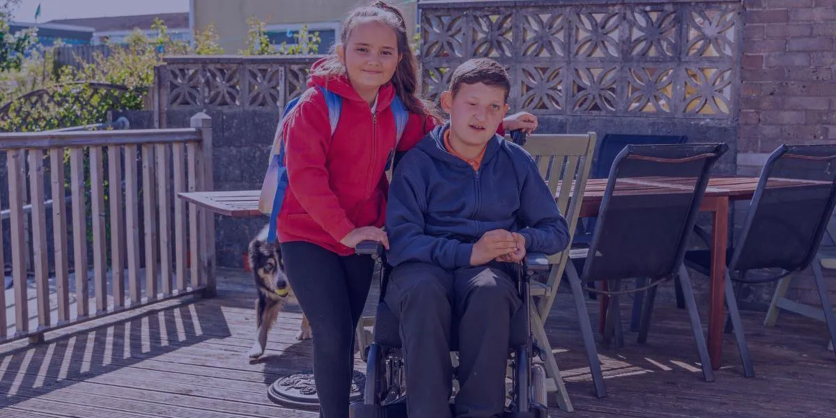 A young girl in a red hoodie standing next to a boy in a wheelchair on a wooden deck, with a dog walking behind them and outdoor furniture in the background.