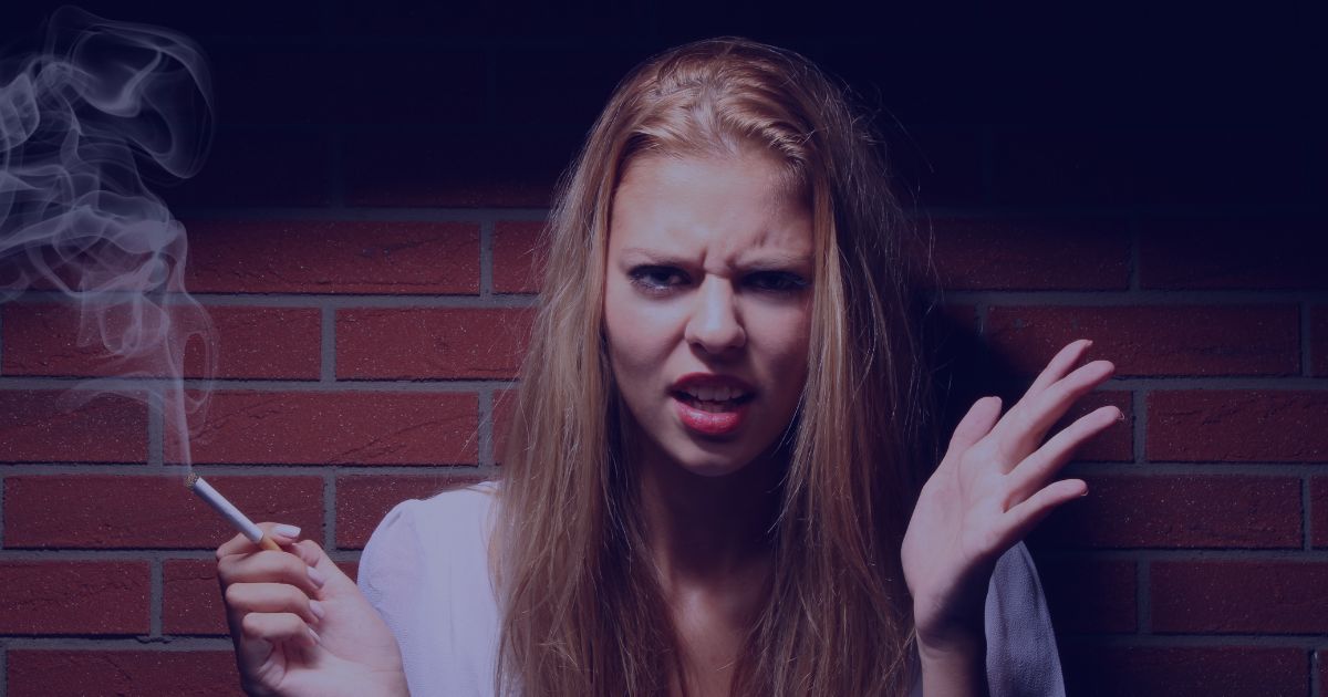 a young female stood against a wall whilst holding a lit cigarette